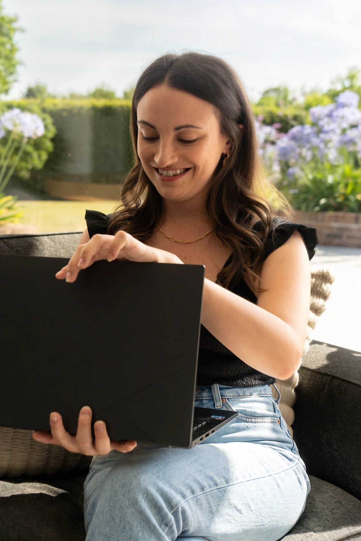 Carly, founder of Nightbloom Studio, smiling while opening a laptop on a sofa, with flowering plantsin the background