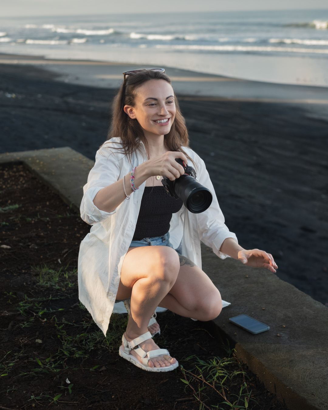 Carly, founder of Nightbloom Studio, crouching and smiling on a beach while holding a camera, black sand and sea behind her
