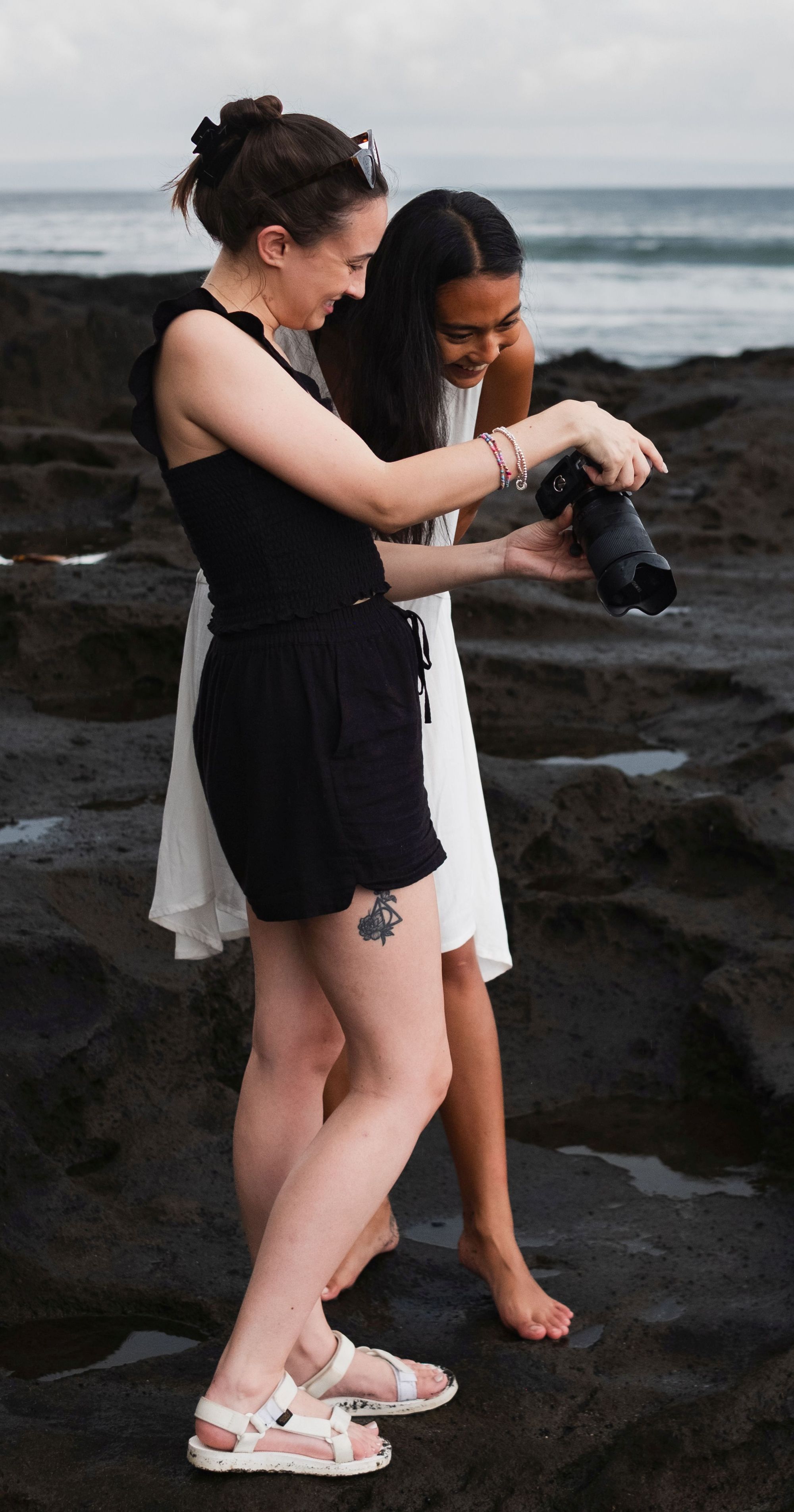 Carly shows a female client photos on her camera on a camera on a rocky coastline. They are both smiling and the sea is behind them.