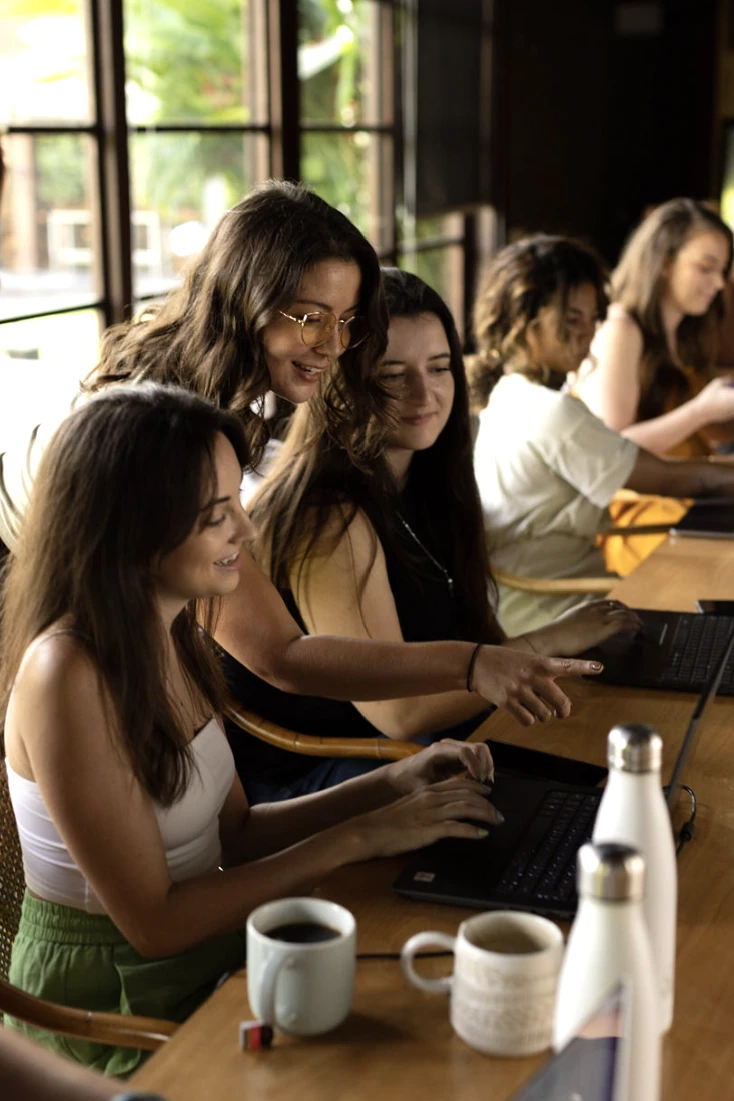 Carly and a group of women smiling and working on laptops together around a table with coffee