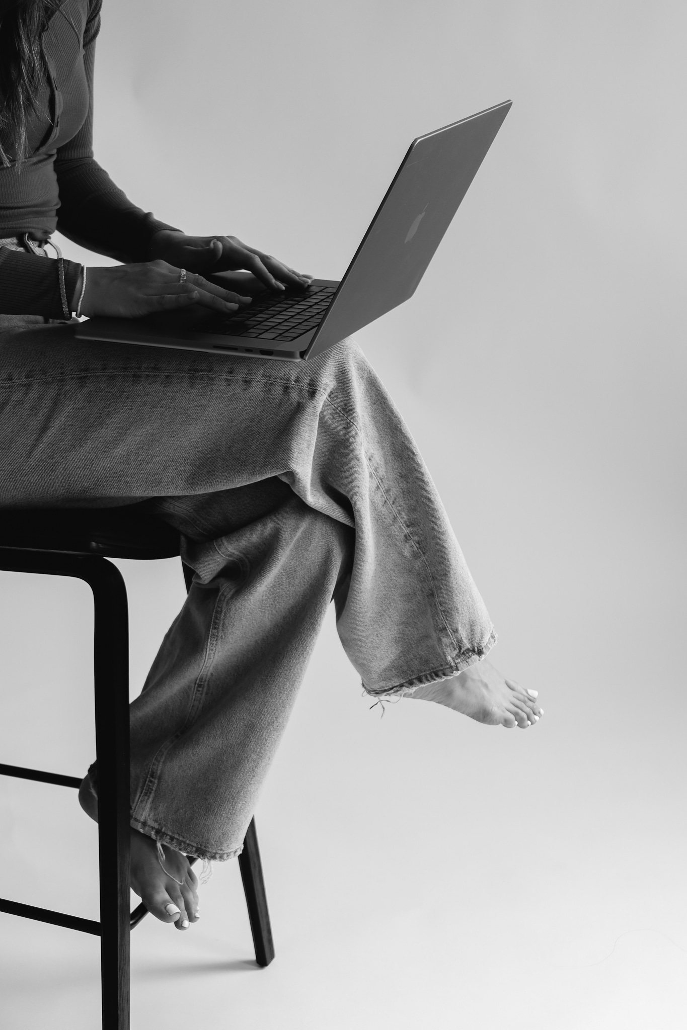 Black and white close-up of hands typing on a laptop balanced on crossed, bare-footed legs (wider shot, foot dangling)